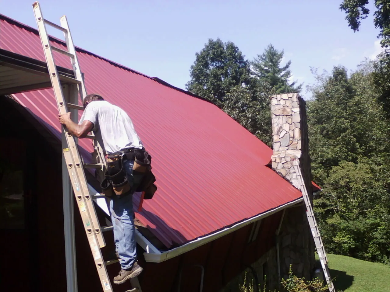 Roofer on a ladder beside a red metal roof and stone chimney — Soffit & Fascia Repair by Roofing Brothers in Shickshinny