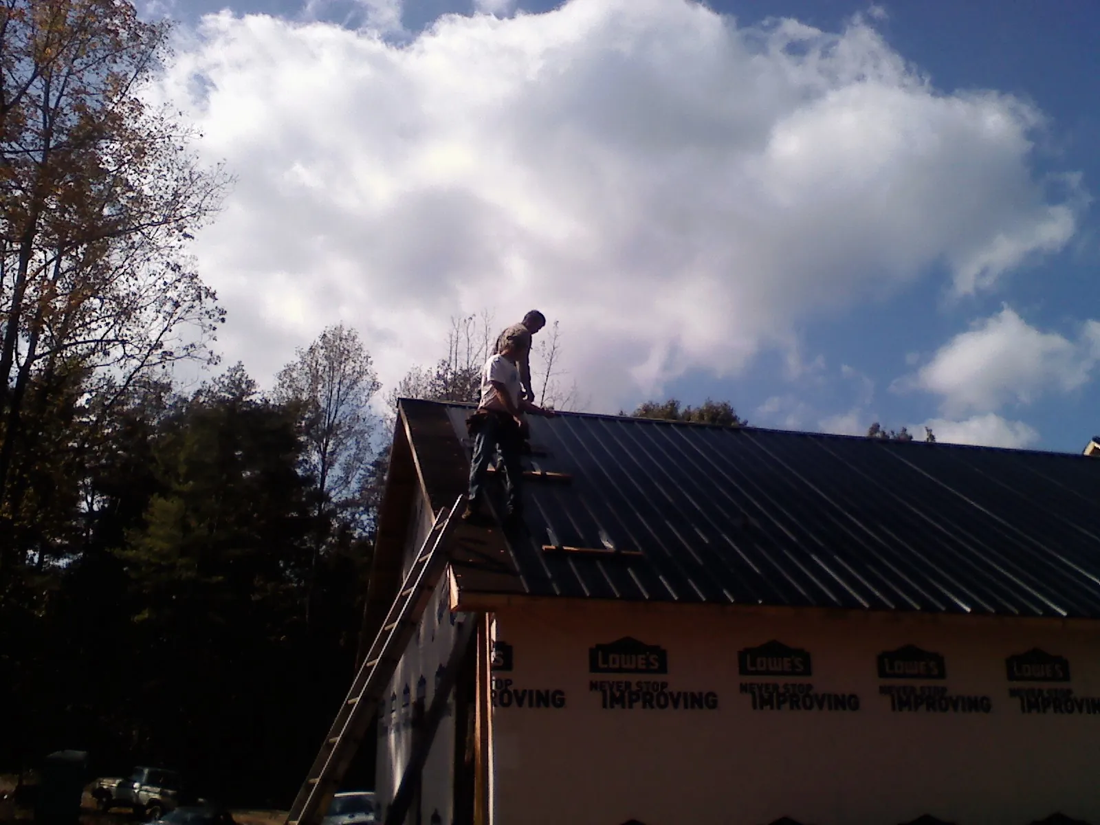 Roofer fastening dark metal panels high against a cloudy sky, from a Roof Inspection job in Shickshinny